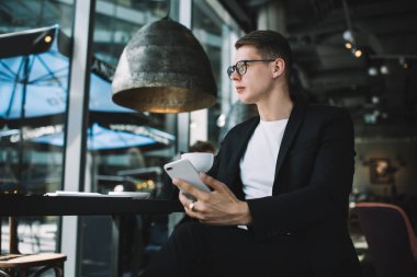 Resmi giyinmiş genç erkek öğrencilerden biri kafede otururken cep telefonu kullanıyor ve başka tarafa bakıyor.