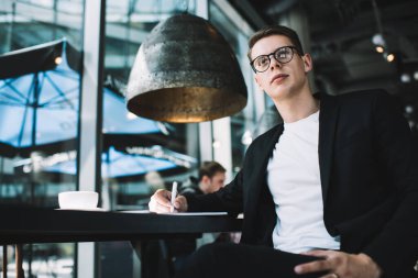 Resmi takım elbiseli ve gözlüklü dalgın erkek yazarın kafede oturmuş gözlerini kaçırırken roman üzerinde çalışmasından.