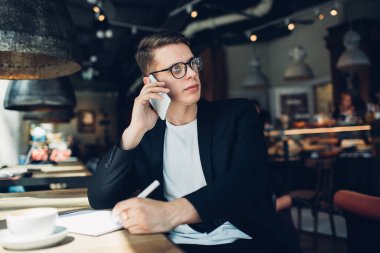 Gözlüklü neşeli akıllı işadamı cep telefonuyla konuşuyor ve karanlık modern kafede, lambanın altında not defterine yazıyor.