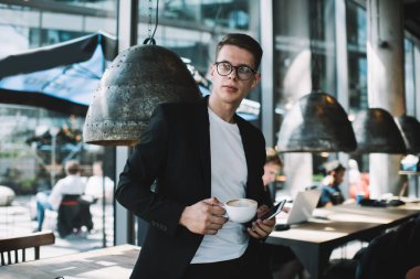 Gözlüklü ve siyah şık takım elbiseli genç girişimci elinde cep telefonu ve kafede sıcak içecekle dikilirken gözlerini kaçırıyor.