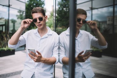 Beyaz gömlekli ve altın yüzüklü ciddi bir adam cep telefonuyla durup güneş gözlüklerine dokunuyor ve gün ışığında şeffaf duvara yaslanıyor.