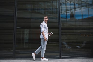 Gündelik kıyafetleri ve beyaz spor ayakkabıları olan bir adamın öğleden sonra cep telefonu ve güneş gözlükleriyle toprak zeminde yürümesi ve geriye bakması.