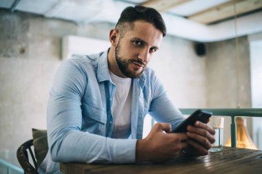 Normal gömlekli ciddi sakallı bir adam modern kafedeki ahşap masada oturuyor ve kameraya bakarken akıllı telefon kullanıyor.