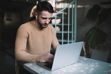 TWS kulaklıklı karizmatik bir adam çevrimiçi çalışıyor ve modern kafede yazı yazarken bilgisayarın monitörüne bakıyor.