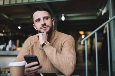 Kafası eğik, bej kazak giymiş ve kol saati takmış, kafede oturmuş çenesine dokunup bakışlarını kaçıran düşünceli genç bir erkek.