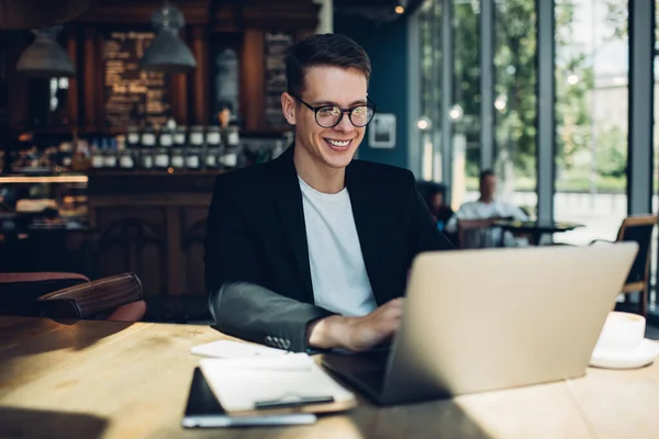 Content male remote worker in formal wear and eyeglasses surfing internet on netbook while sitting at table with organizer and cup of coffee at cafeteria