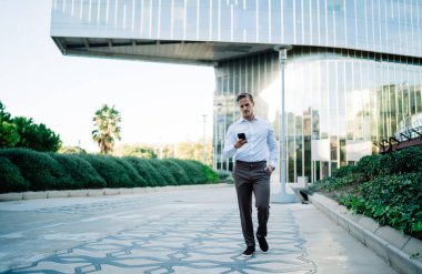 Gündüz vakti iş yerinden çıkarken telefonda mesajlaşan, resmi giyinmiş, odaklanmış genç bir adam vücudu.