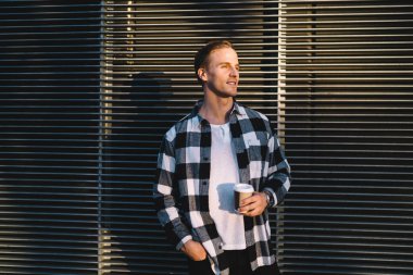 Yakışıklı, modaya uygun kıyafetler giymiş beyaz adam kameraya bakıyor. 20 'li yaşlarda, kahve fincanı taşıyan genç bir erkek sokak ortamlarında gülümsüyor.