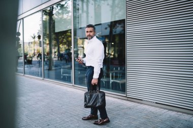 Modern akıllı telefon cihazında 4G internet bağlantısı kullanan beyaz tişörtlü beyaz tenli tam boy bir işadamı ve cep telefonu ve moda çantalı orta yaşlı bir işadamı kameraya bakıyor.