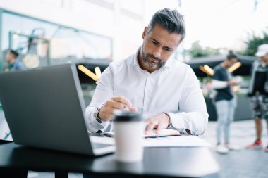 Profesyonel Kafkasyalı yönetici, uzaktaki bir kafede kişisel planlamacı kullanıyor. Modern laptop teknolojisi ile masada oturuyor ve bilgi notlarını analiz ediyor. Beyaz gömlekli iş adamı çalışıyor.