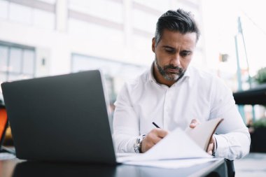 Verimli bir strateji planlarken bilgilendirici notlar alan ciddi yetişkin işadamı, sokak kafede uzaktan çalışan beyaz erkek çalışanı araştırma için dizüstü bilgisayarlı teknolojiyle masada oturuyor.