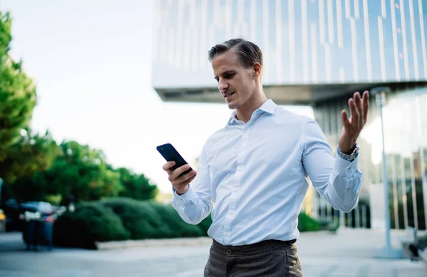 年轻的男性高管一边与市中心的现代摩天大楼相对照 一边挥手表示不满 一边在电话上查看电子邮件 — 图库照片