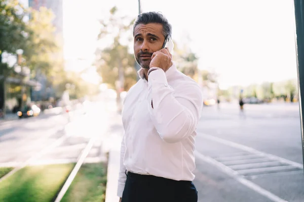 Handsome entrepreneur 40 years old calling to service for consultancy puzzled on received information, serious businessman in white shirt making international smartphone conversation in roaming