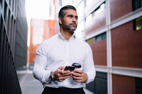 Pensive businessman in white shirt pondering on received mobile newsletter standing at urbanity with takeaway cup, contemplative male entrepreneur with cellphone and coffee to go in hand thinking
