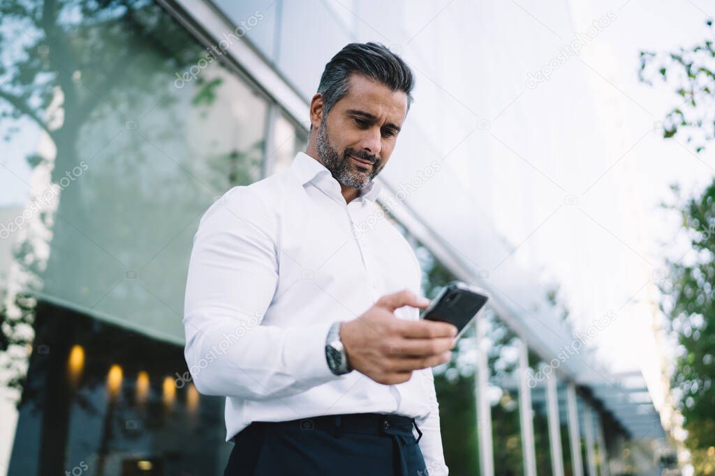 Below view of mature lawyer with stubble using mobile application for chatting in business chat, Caucasian male proud CEO in white shirt searching banking app for making online payment and banking