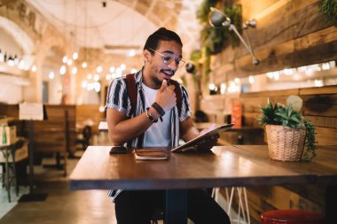 Gözlüklü koyu tenli bir erkek dijital tablet kullanarak online kitap okuyor. 20 'li yaşlarda Afro-Amerikan hipster bir genç. İnternette 4G bağlantısı kursundan memnun.