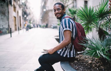 Mutlu etnik erkek kitap tutuyor ve boş bir sokakta çiçek tarhında oturmuş gülümsüyor ve kameraya bakıyor.