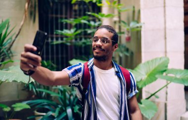 Modayı takip eden yaz kıyafetleri ve sırt çantalı gözlüklü, akıllı telefonlu, gündüz vakti sokakta yürürken selfie çeken pozitif sakallı bir etnik adam.