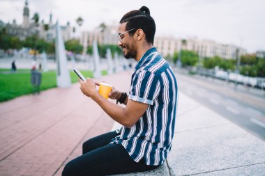 Gülümseyen Afro-Amerikalı genç adam akıllı telefondan mesajları kontrol ediyor 4G internet kullanıyor, pozitif esmer tenli hipster şehirde arkadaşlarıyla çevrimiçi iletişim kuruyor.