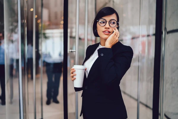 カフェに立ちスマートフォンで話すテイクアウトコーヒーのカップと正式な摩耗や眼鏡で自信のある民族経営の女性 — ストック写真