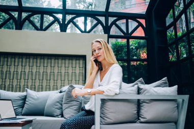 Resmi kıyafetli iyimser kadın girişimcinin cep telefonuyla konuşurken masanın yanındaki rahat koltukta dizüstü bilgisayarla kameraya bakmasından.