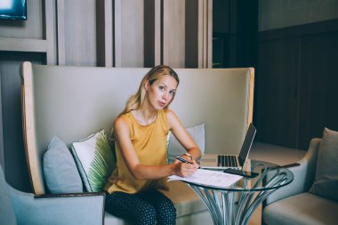 Rahat kadın girişimci, günlük kıyafetlerle dokümanları dolduruyor ve kafede dizüstü bilgisayarla oturup kameraya bakarken uzaktan çalışıyor.