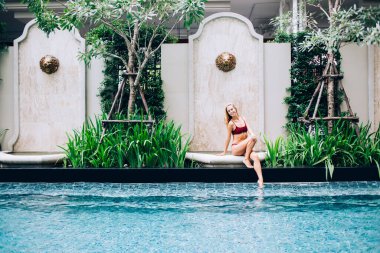 Mayo giymiş cılız bir kadın havuz kenarında dinleniyor, ayaklarıyla suya giriyor ve tatil boyunca dinleniyor ve kameraya bakıyor.