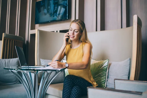 穿着雅致服装的年轻女自由撰稿人一边阅读财务报告 一边在智能手机上讲话 一边在咖啡店的桌上与笔记本电脑远程合作 — 图库照片