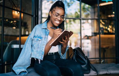 Günlük kıyafetli mutlu siyahi bayan, kafenin cam duvarına karşı otururken not defterine not alıyor ve dişlek gülümsüyor.