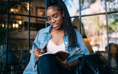 Not defterinde çizim yapan güzel koyu renk tenli zeki bir öğrenci. Hobisiyle vakit geçirmekten zevk alıyor. Koyu tenli olumlu bir kız öğrenci kafenin içinde not defteriyle notlar alıyor.