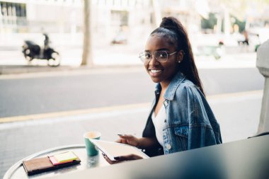 Kot ceketli, uzun siyah saçlı, kameraya gülümseyen ve tek kullanımlık kahveyle sokak kafesinde oturan genç bir kadının yan görüntüsü.