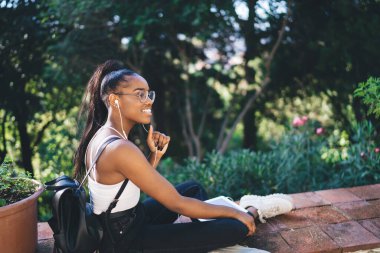 Yazlık kıyafetleri ve gözlükleriyle parkta günün tadını çıkarırken tabletten müzik dinleyen çekici siyah genç kız.
