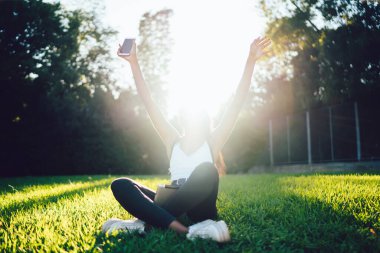Nilüfer çiçeği pozunda otururken ve parktaki güneşli bir günde el kaldırırken gündelik kıyafetleriyle tanınmayan bir kadın.