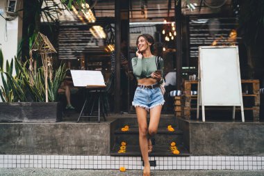 Güneşli bir günde sokakta yürürken gülümseyen ve bakışlarını kaçıran günlük yaz kıyafetiyle uzun boylu, iyimser bir kadın.