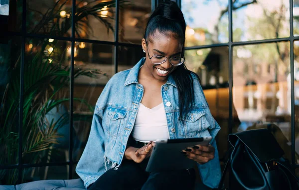 Cheerful dark skinned woman in eyewear for vision correction smiling checking salles in web store on digital tablet, positive african american hipster girl share media and multimedia content online