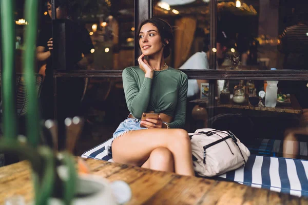 穿着休闲装的多愁善感的女人坐在舒适的长椅上 一边在咖啡店休息 一边沉思地回头看 — 图库照片