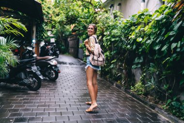 Yeşil bitkilerin yanında asfalt yolda yürürken gülümseyen ve bakışlarını çeviren günlük kıyafetli neşeli kadın.