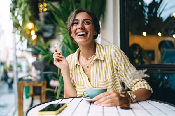 カジュアルな服装の正の若い女性が笑顔とコーヒーカップを飲みながら 屋外カフェテリアで時間を過ごしながらティースプーンを保持 — ストック写真