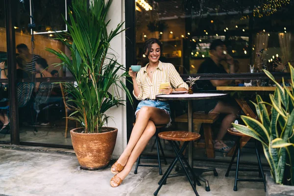 Happy young female in summer outfit with cup of coffee smiling and browsing mobile phone while resting outside of cozy restaurant at daytime