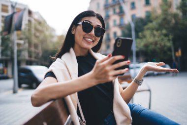 Neşeli bin yıllık kadın blogcu güneş gözlüklü sosyal ağlarda blog haberlerini kontrol ediyor kentteki bankta oturmuş gülümseyen Asyalı kadın 4G ile cep telefonu ve kısa mesaj yolluyor.