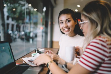 Kameraya bakan neşeli Asyalı kadın portresi Kafe 'deki uzak iş için Kafkasyalı meslektaşıyla boş zaman geçiriyor, pozitif hipster kız birlikte zaman geçiriyor iletişim kuruyor ve cihazlar kullanıyor.