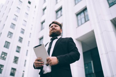 Kendine güvenen sakallı, zarif takım elbiseli, elinde tabletle modern yüksek katlı iş merkezinin yanında duran iş adamlarının liderinden.