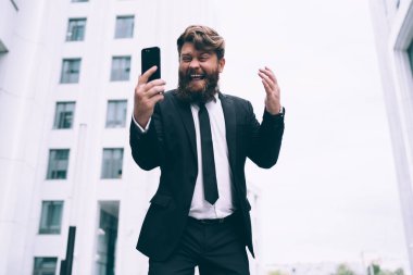 Resmi takım elbiseli mutlu sakallı adam gündüz vakti sokakta dikiliyor ve gülerken ve ekrana bakarken akıllı telefon kullanıyor.