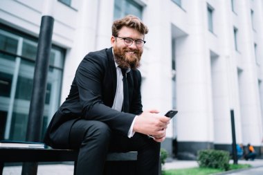 Resmi takım elbiseli neşeli bir genç adam. Ofis binasının bahçesindeki bankta dinleniyor ve parlak bir gülümsemeyle kameraya bakıyor.