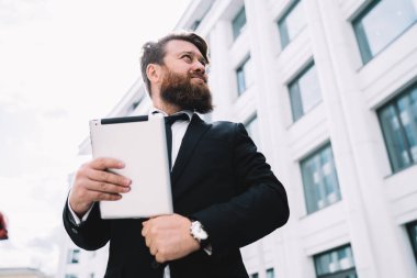 Düşük açılı sakallı adam elinde aletle işten çıkarken modern binanın yanında duruyor.