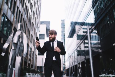 Gülümseyen sakallı, resmi takım elbiseli ve gür sakallı bir çalışanın modern gökdelenler ve akıllı telefonlara bakarken gülümsemesi arasında şehirde durmasından.