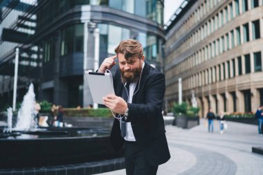 Resmi takım elbiseli genç ciddi işadamı, şehir parkında fıskiyeli ve sokak kafeli bir tabletle video araması yaparken çalışanı azarlıyor.
