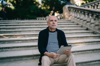Siyah ceketli ve çizgili gömlekli düşünceli bir erkek parktaki merdivenlerde dinleniyor ve molada tablet okurken rüya görüyor.