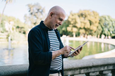 Sıradan giyinmiş, taş tırabzanların yanında duran ve parkta yürürken tablet üzerinde sohbet eden yaşlı adamın yan görüntüsü.
