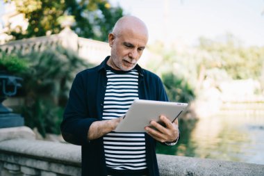 Gri sakallı ve kel kafalı ciddi yaşlı bir adam şehir parkında dinlenirken taş parmaklıkların yanında durup tablet karıştırıyor.